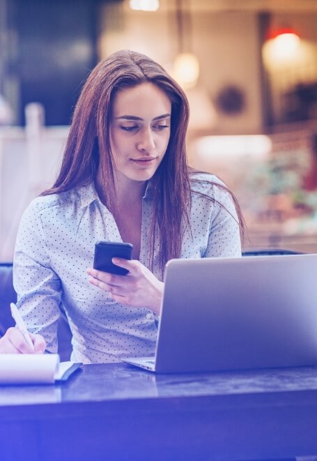 Foto de uma mulher segurando um celular e olhando para a tela de um laptop